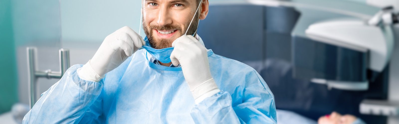 Ein Augenchirurg in Op-Kleidung zieht seinen Mundschutz hoch. Hinter ihm sieht man unscharf eine Patientin unter einem Laser.